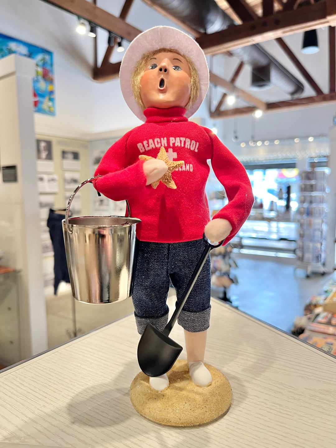 Figurine of a person in a red 'Beach Patrol' shirt holding a shovel and bucket, standing on a wooden surface.