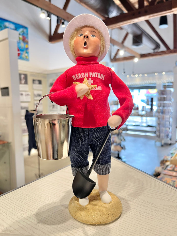 Figurine of a person in a red 'Beach Patrol' shirt holding a shovel and bucket, standing on a wooden surface.