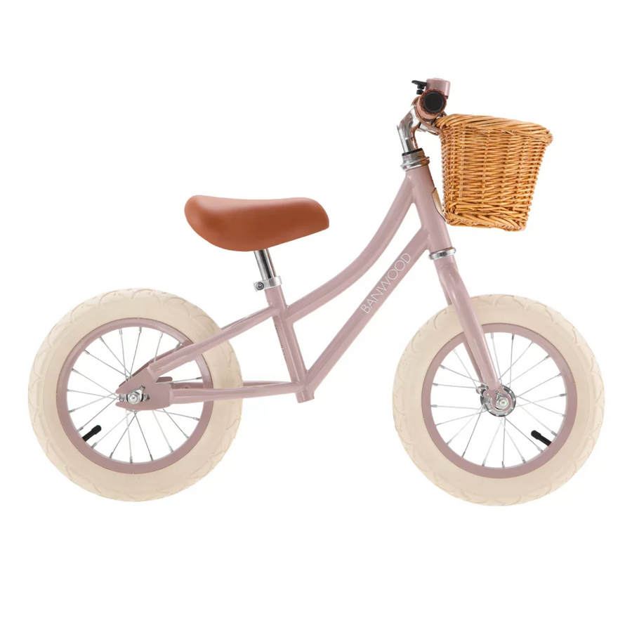 Pink balance bike with a brown seat and wicker basket on a white background