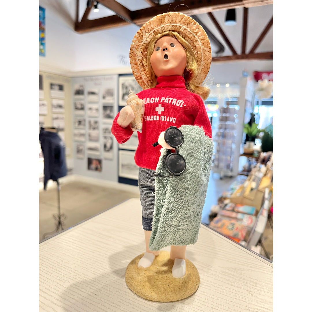 Decorative figure of a woman in a red shirt and straw hat holding sunglasses, standing on a wooden surface.