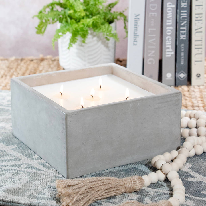 Square concrete candle holder with lit candles on a textured surface with books and a plant in the background.