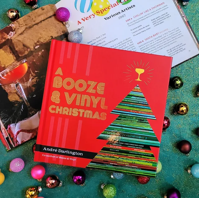 Book titled 'A Booze & Vinyl Christmas' by André Darlington surrounded by Christmas decorations on a green surface.